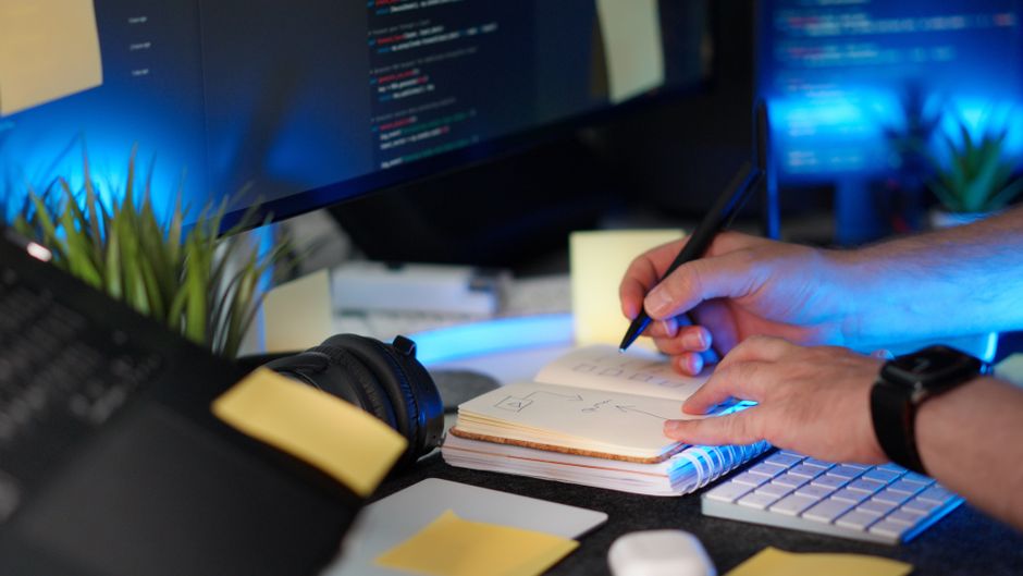 A person planning a software project with handwritten notes and code on a monitor in a modern workspace.